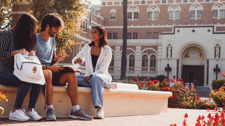 Three students sit on the fountain outside of Doheny Library discussing a book as the sun sets.