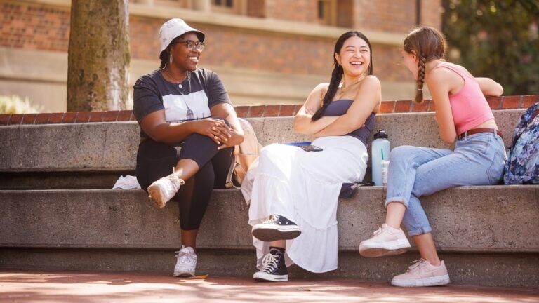 Three students sit and laugh on bench on a sunny day. Pane Image 1