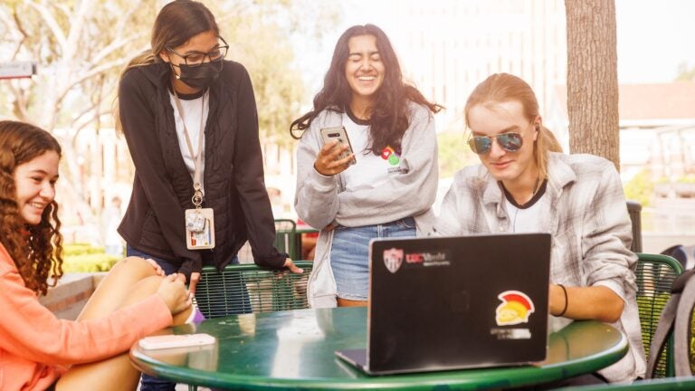 Four students laugh at a computer screen at a table outside the USC Everybody