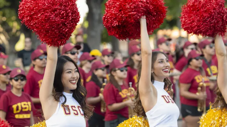 Two USC Song Girls cheerfully hold up red pom-poms with the Trojan Marching band in the back. Pane Image 1