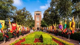 USC graduates carrying school banners during a commencement procession in front of Bovard Auditorium.